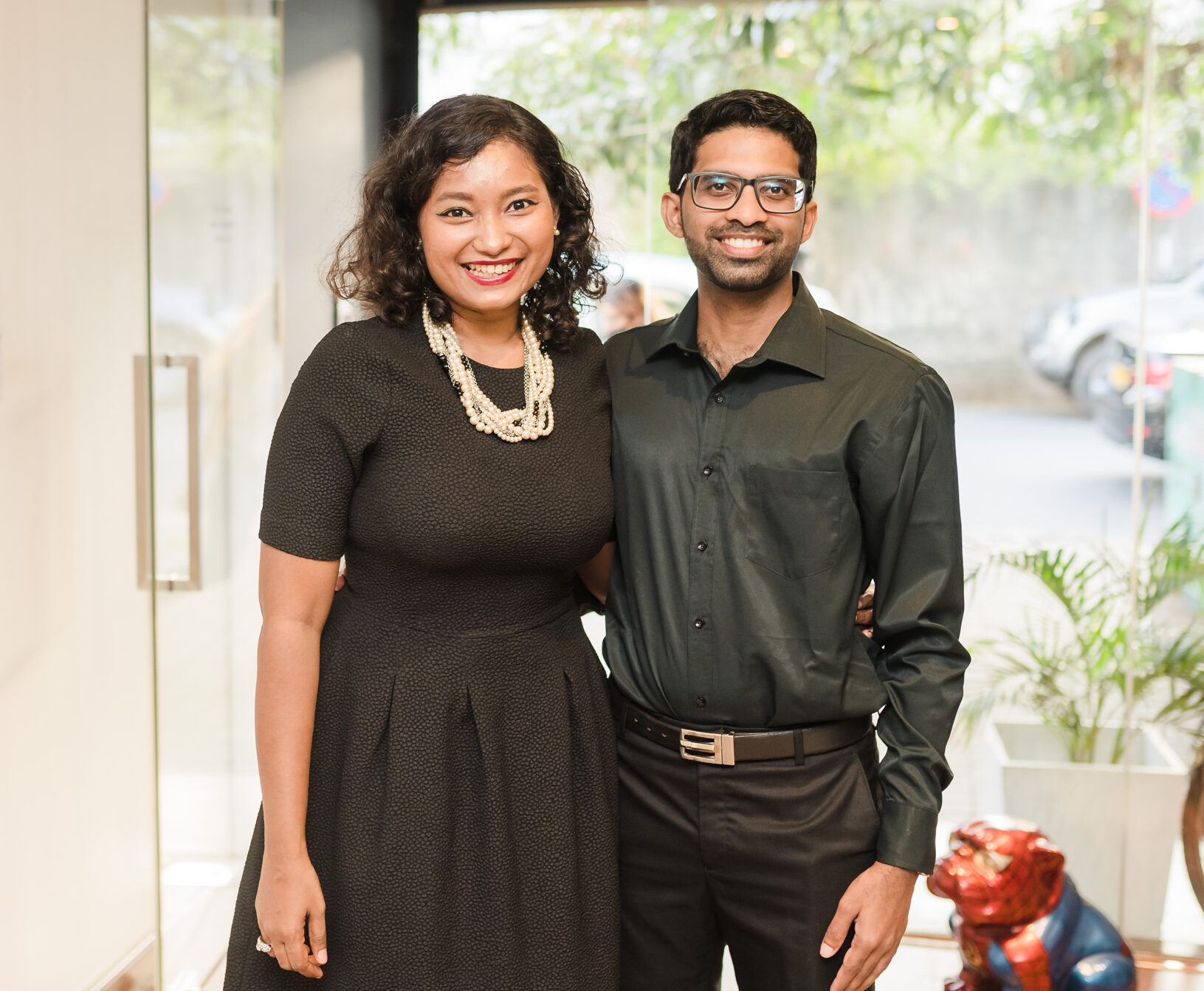 A young woman in a black dress and a young man in a black shirt pose for a photograph with glass doors in the background for Mandy Projects launch